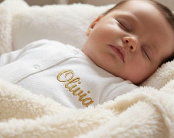 Könnte beinhalten: Ein schlafendes Baby in einem weißen Strampler mit dem Namen "Olivia" in Gold gestickt. Das Baby liegt in einer weichen, cremefarbenen Decke.