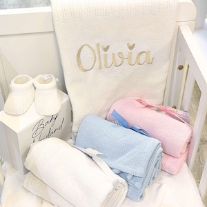 May include: A collection of baby items, including a white blanket embroidered with the name "Olivia" in gold, folded blankets in blue and pink, and a pair of white baby booties. The items are arranged in a white crib.