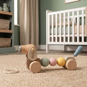 May include: A wooden pull-along toy shaped like a dog with a natural wood finish, featuring a light blue tail, a yellow bead, a pink bead, and a sage green bead. The dog's ear is personalised with the name SOPHIE in black text and includes a white cord.