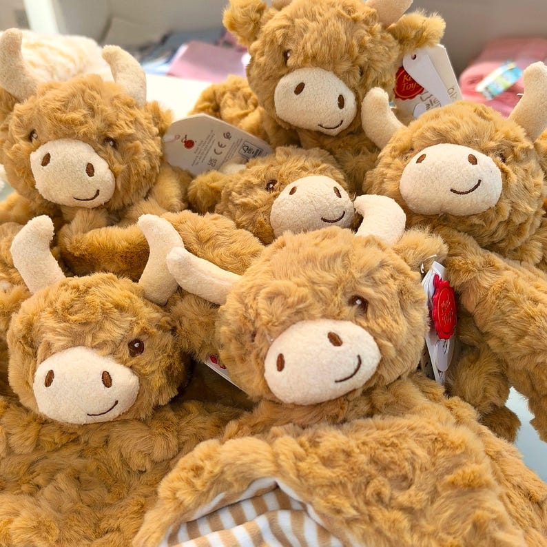 May include: A collection of plush Highland cow stuffed animals. Each cow has a light brown, shaggy fur body, a white face with a smiling expression, and cream-colored horns. Some have tags attached.