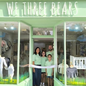 K&ouml;nnte beinhalten: Ein Gesch&auml;ft mit der Aufschrift "WE THREE BEARS" in wei&szlig;en Buchstaben &uuml;ber dem Eingang. Eine vierk&ouml;pfige Familie steht im T&uuml;rrahmen und schneidet ein Band durch. Im Inneren des Gesch&auml;fts werden Kinderkleidung ausgestellt.