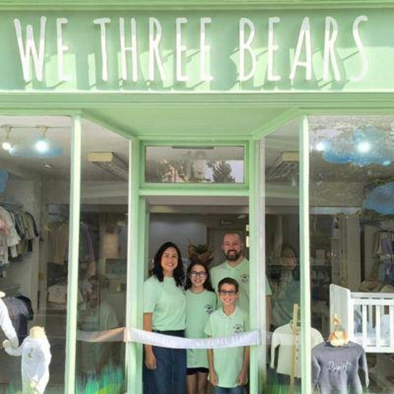 May include: A family stands in front of a store with the text "WE THREE BEARS" above the entrance. The store is painted a light green color. The family is wearing matching light green shirts. A white ribbon is stretched across the doorway.