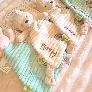 May include: Soft, plush Winnie the Pooh themed baby items. The image shows a cream-colored bear and several corduroy-textured baby comfort items in cream and mint green. The names "Maria" and "Noah" are embroidered on the items.