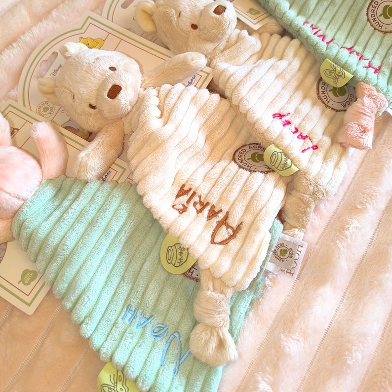 May include: Soft, plush Winnie the Pooh themed baby items. The image shows a cream-colored bear and several corduroy-textured baby comfort items in cream and mint green. The names "Maria" and "Noah" are embroidered on the items.