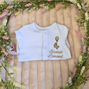 K&ouml;nnte beinhalten: Wei&szlig;er Baby-Body mit Knopfleiste. Der Body hat ein gesticktes Design eines B&auml;ren, der einen Ballon h&auml;lt, und den Namen "Ernest Lomond" in Goldfaden. Der Body ist von einem Blumengesteck umgeben.