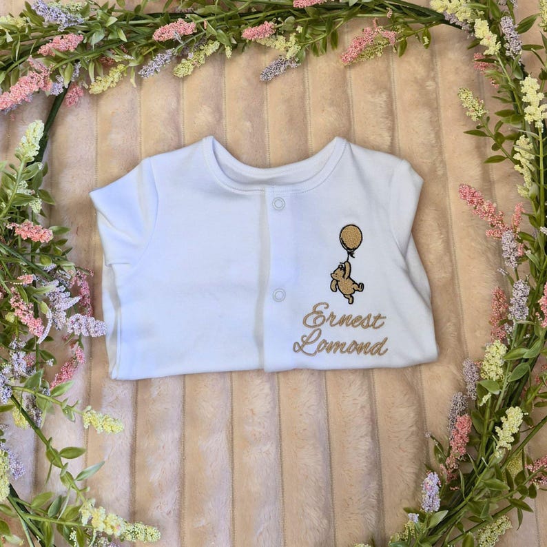 K&ouml;nnte beinhalten: Wei&szlig;er Baby-Body mit Knopfleiste. Der Body hat ein gesticktes Design eines B&auml;ren, der einen Ballon h&auml;lt, und den Namen "Ernest Lomond" in Goldfaden. Der Body ist von einem Blumengesteck umgeben.