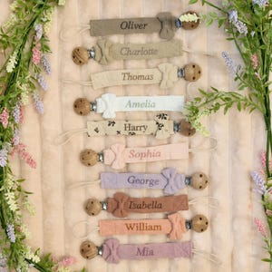 May include: A selection of personalised baby dummy clips in pastel shades, each embroidered with a name. The clips have wooden beads and a bow, displayed on a soft, textured background, with floral accents.