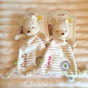 May include: Two Winnie the Pooh security blankets. Each plush toy has a cream-colored body with a corduroy texture and a Winnie the Pooh head. The blankets have the names "Maria" and "Lacey" embroidered on them, along with the "Hundred Acre Wood" logo.