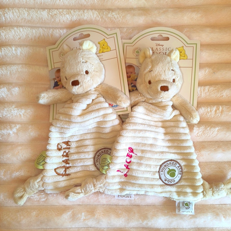 May include: Two Winnie the Pooh security blankets. Each plush toy has a cream-colored body with a corduroy texture and a Winnie the Pooh head. The blankets have the names "Maria" and "Lacey" embroidered on them, along with the "Hundred Acre Wood" logo.