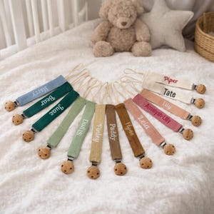May include: A selection of personalised baby dummy clips in shades of blue, green, beige, and pink. Each clip has a wooden bead and is embroidered with a name in a matching colour. The clips are arranged in a circular pattern.