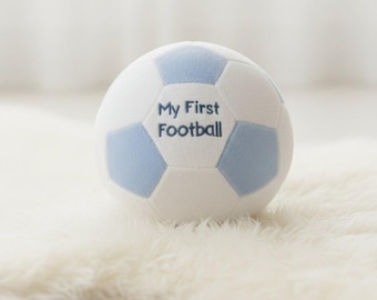 Könnte beinhalten: Ein Plüsch-Fußball mit einem klassischen Fünfeck-Muster in Weiß und Hellblau. Der Ball ist mit dem dunkelblauen Text My First Football bestickt, der auf einem der weißen Felder zentriert ist, was ein weiches und haptisches Sportspielzeug ergibt.