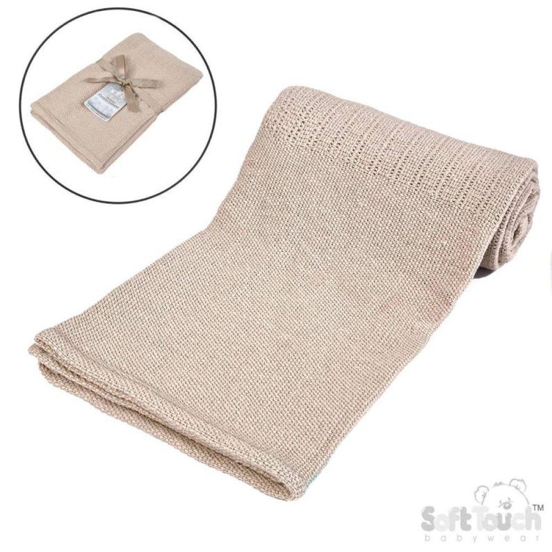 May include: A beige knitted baby blanket, partially rolled, with a folded section at the bottom. A smaller, folded blanket with a tag and ribbon is in a circular inset. The blanket is made of a soft, breathable material.
