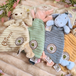 May include: Four plush baby comforters featuring Winnie the Pooh characters. Each comforter has a character head and a corduroy body in cream, green, blue, and yellow. The comforters have the text "Hundred Acre Wood" and character names.