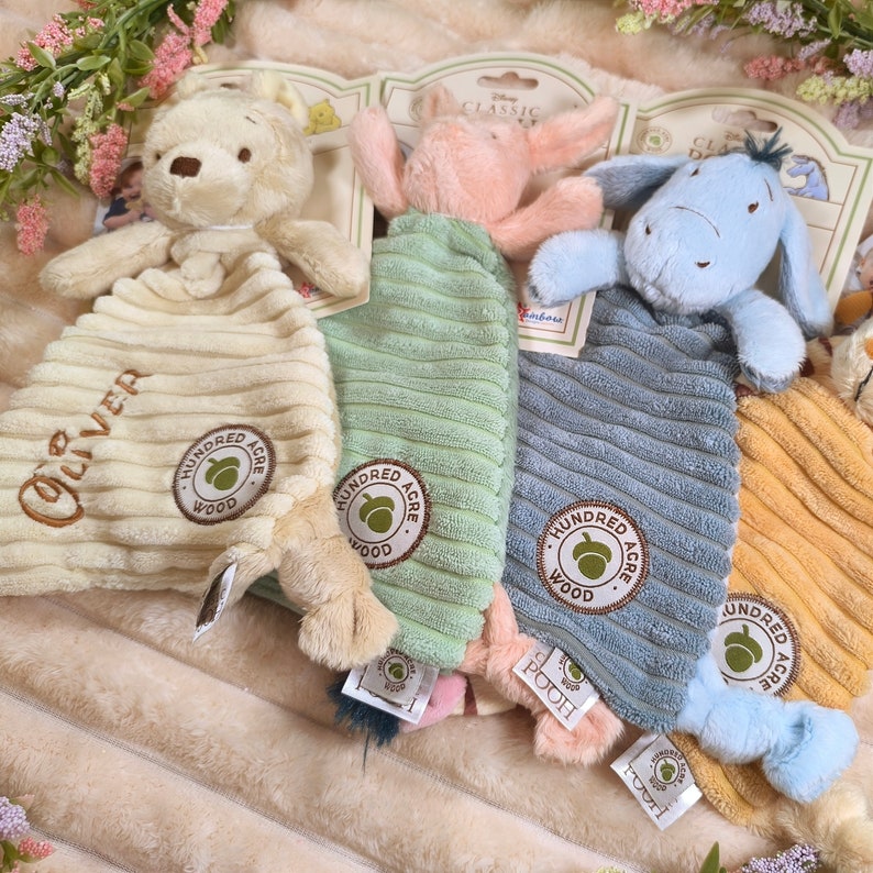 May include: Four plush baby comforters featuring Winnie the Pooh characters. Each comforter has a character head and a corduroy body in cream, green, blue, and yellow. The comforters have the text "Hundred Acre Wood" and character names.