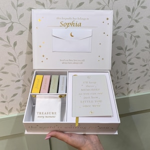 May include: A white keepsake box with gold detailing, personalised with the name "Sophia." Inside are colourful keepsake cards, a small pouch with the words "TREASURE every moment," and a card with a poem. The box is designed to hold baby keepsakes.