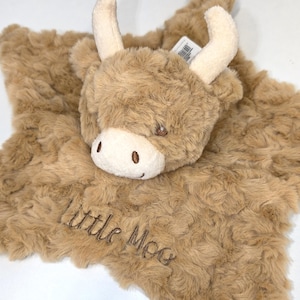 May include: A plush, brown Highland cow security blanket with a soft, textured surface. The cow has cream-colored horns, a white face, and embroidered details. The words "Little Moo" are embroidered in brown on the blanket.