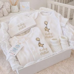 K&ouml;nnte beinhalten: Ein wei&szlig;es Baby-Geschenkset in einer wei&szlig;en Box. Das Set enth&auml;lt einen Hut, zwei Bodys und gestrickte Babyschuhe. Jeder Artikel ist mit dem Namen "Baby Millar" in Gold und einem Cartoon-B&auml;ren mit einem Ballon bestickt.
