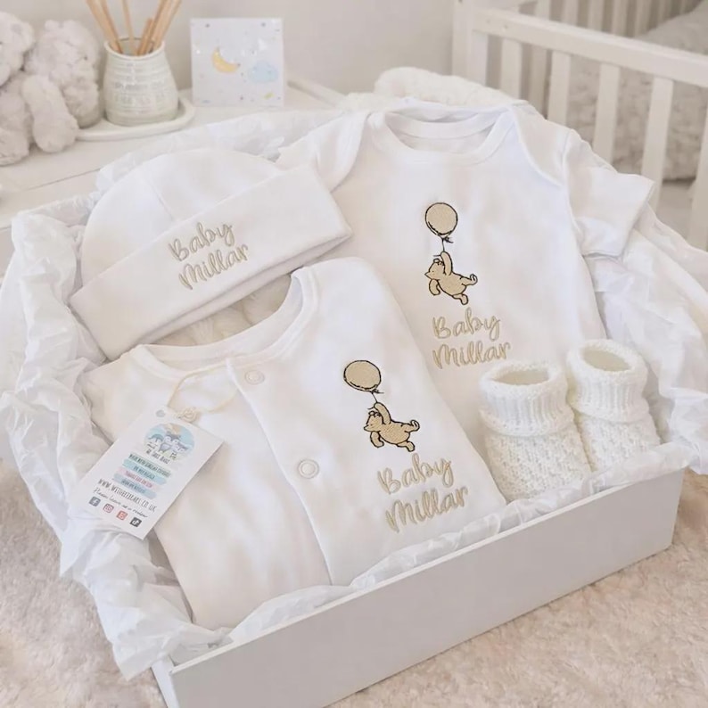 K&ouml;nnte beinhalten: Ein wei&szlig;es Baby-Geschenkset in einer wei&szlig;en Box. Das Set enth&auml;lt einen Hut, zwei Bodys und gestrickte Babyschuhe. Jeder Artikel ist mit dem Namen "Baby Millar" in Gold und einem Cartoon-B&auml;ren mit einem Ballon bestickt.
