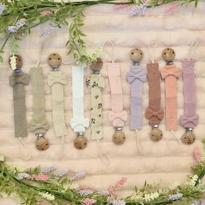 May include: A selection of baby pacifier clips in shades of beige, white, pink, and purple. Each clip has a wooden bead, a fabric strap, and a bow. The clips are displayed on a soft, textured surface, with floral decorations around them.