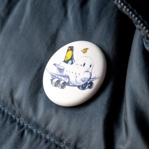 Könnte beinhalten: Ein weißer Ansteckbutton mit einem Cartoon-Flugzeug-Design. Das Flugzeug hat ein lächelndes Gesicht, Flügel und Räder. Das Heck ist gelb, schwarz und grün. Über dem Flugzeug befindet sich ein kleines gelbes Insekt. Der Button befindet sich auf einer dunkelblauen Jacke.