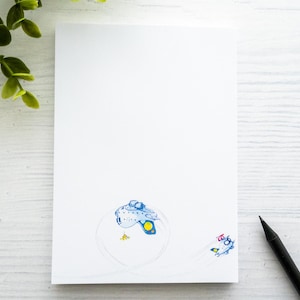 Könnte beinhalten: Eine weiße leere Karte mit einer blauen und gelben Illustration eines Fisches in einem Kreis. Ein schwarzer Bleistift liegt auf einer weißen Holzoberfläche.