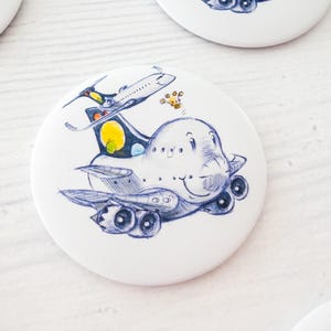 Könnte beinhalten: Weiße Keramikuntersetzer mit einem skurrilen Flugzeugdesign. Die Illustration zeigt ein lächelndes Flugzeug mit einem kleineren Flugzeug darüber, alles in blauer Tinte. Eine Giraffe ist ebenfalls enthalten.