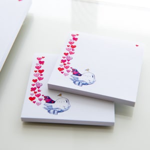 Könnte beinhalten: Zwei weiße Notizblöcke mit einem rosa Herzrand und einer Illustration eines blauen Vogels. Der Vogel sitzt auf einer Wolke mit einem kleinen gelben Flugzeug, das darüber fliegt.