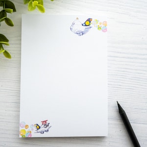 Könnte beinhalten: Ein weißer Notizblock mit einem bunten Vogel- und Blasendesign. Der Notizblock hat eine leere Seite mit einem schwarzen Bleistift auf einer weißen Holzoberfläche.