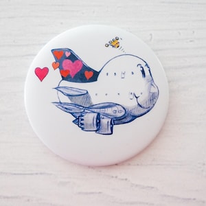 Könnte beinhalten: Ein weißer Button mit einer skurrilen Illustration eines Flugzeugs. Das Flugzeug hat ein lächelndes Gesicht, herzförmige Akzente am Heck und eine kleine Biene. Das Design ist in Blau, Rot und Rosa gehalten.