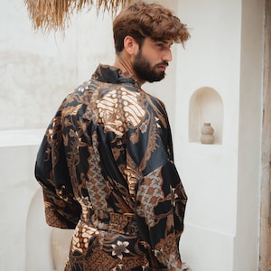 May include: A black robe with a brown and white batik pattern. The robe is worn by a person standing in front of a white wall. The robe has a belt and long sleeves. The background includes a thatched roof and a small alcove.