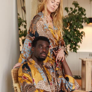 May include: A couple wearing matching patterned silk satin kimono robes in shades of yellow, blue, and brown. The robes feature an intricate design. One person is seated in a wicker chair, while the other stands behind them.