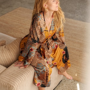 K&ouml;nnte beinhalten: Ein gemusterter Jumpsuit in Senfgelb, T&uuml;rkis und Schwarz. Das Kleidungsst&uuml;ck hat ein Oberteil im Wickelstil und eine Hose mit weitem Bein. Das Model tr&auml;gt goldene Creolen und eine Halskette. Der Jumpsuit wird mit einem passenden Stoffg&uuml;rtel in der Taille gebunden.