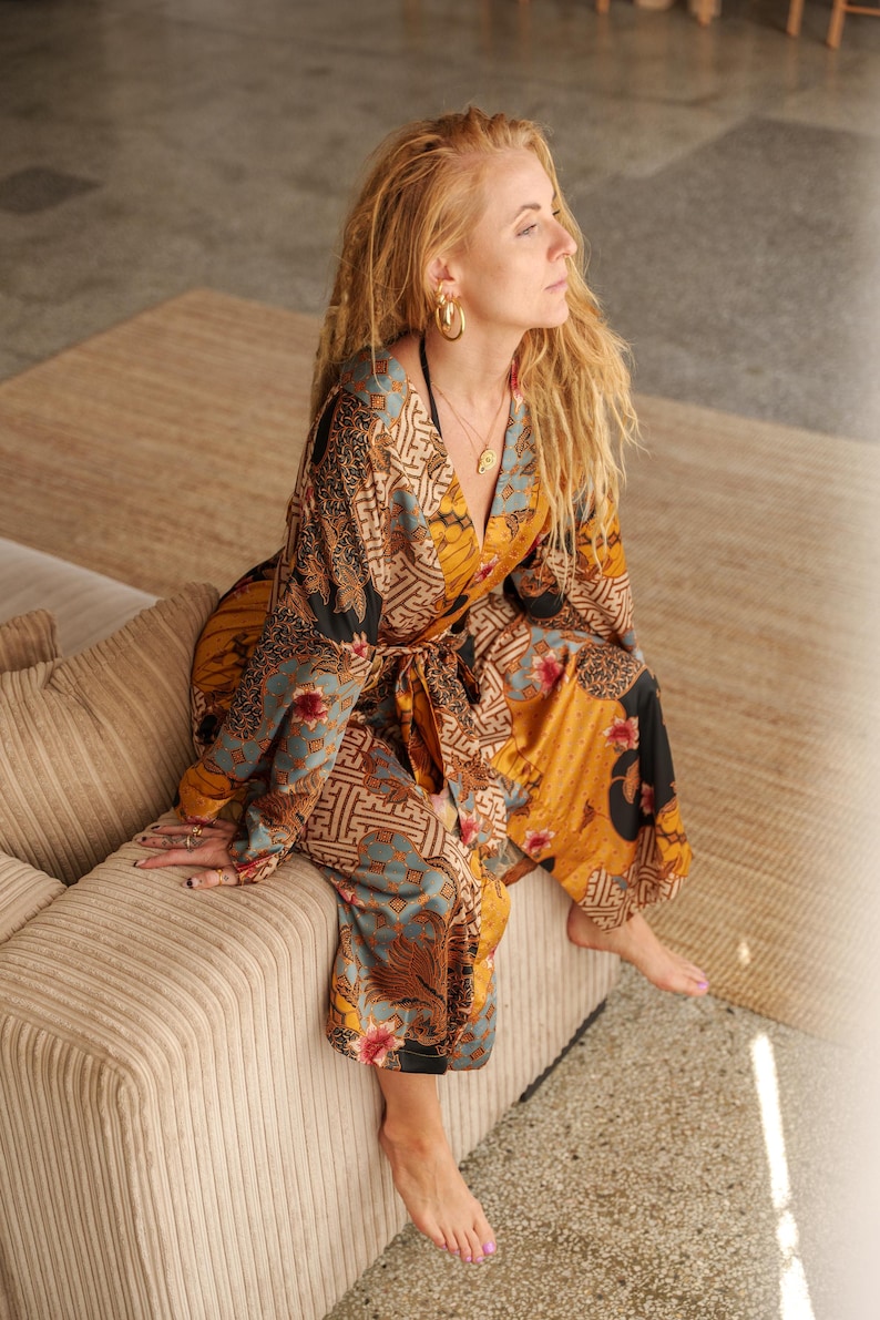 K&ouml;nnte beinhalten: Ein gemusterter Jumpsuit in Senfgelb, T&uuml;rkis und Schwarz. Das Kleidungsst&uuml;ck hat ein Oberteil im Wickelstil und eine Hose mit weitem Bein. Das Model tr&auml;gt goldene Creolen und eine Halskette. Der Jumpsuit wird mit einem passenden Stoffg&uuml;rtel in der Taille gebunden.