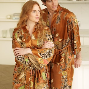 Puede incluir: Albornoces de seda marrones a juego con un estampado floral y geométrico. Los albornoces tienen mangas largas y se atan a la cintura. Los albornoces son de color marrón cálido con toques de verde, rojo y blanco.