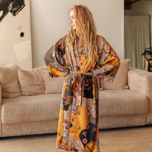 May include: A long, patterned satin silk kimono robe in shades of gold, brown, black, and blue. The robe features a belt and a patchwork design with floral and geometric elements. The model is standing in a room with a beige sofa and a woven rug.