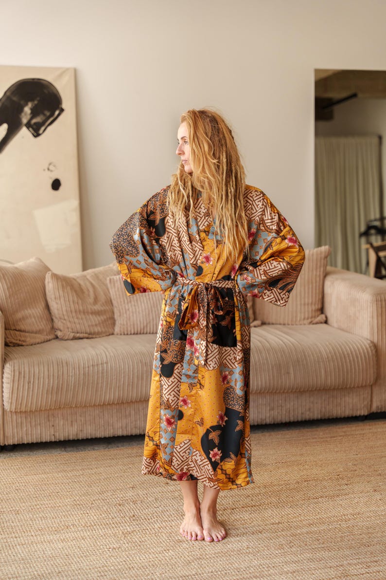 May include: A long, patterned satin silk kimono robe in shades of gold, brown, black, and blue. The robe features a belt and a patchwork design with floral and geometric elements. The model is standing in a room with a beige sofa and a woven rug.