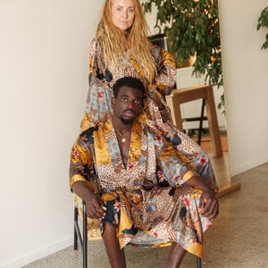 May include: Two people wearing matching satin silk kimono patterned robes. The robes feature a mix of colours including gold, blue, and brown. The robes have a floral and geometric design. One person is seated in a chair, while the other stands behind them.