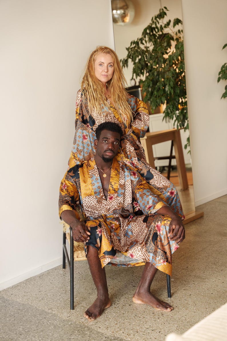 May include: Two people wearing matching satin silk kimono patterned robes. The robes feature a mix of colours including gold, blue, and brown. The robes have a floral and geometric design. One person is seated in a chair, while the other stands behind them.