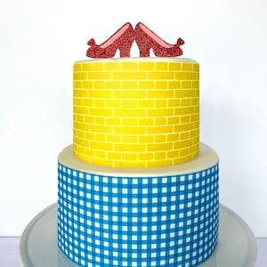May include: A two-tiered cake decorated with a yellow brick road pattern on the top tier and a blue and white gingham pattern on the bottom tier. The cake is topped with a pair of red glitter ruby slippers.