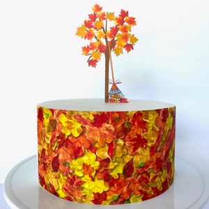 May include: A round cake decorated with a fall leaf pattern. A paper cake topper features a tree with orange and yellow leaves and a rake.