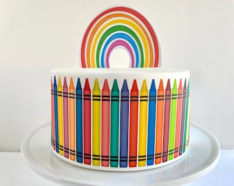 May include: A white cake decorated with a rainbow and a border of colorful crayons. The cake is on a white cake stand.