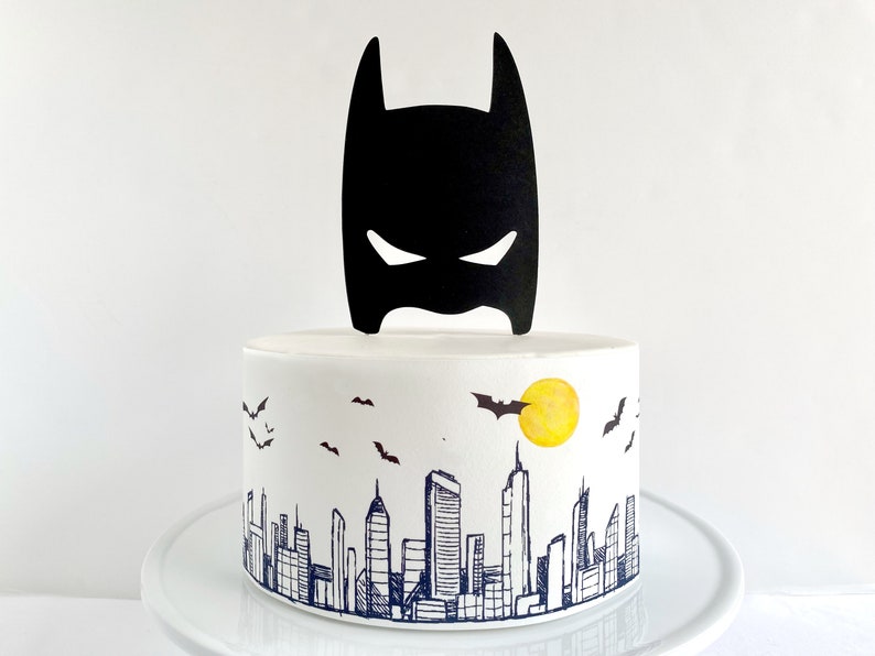 Puede incluir: Un pastel blanco decorado con un adorno de m&aacute;scara de Batman negro y un dise&ntilde;o de ciudad con una luna amarilla y murci&eacute;lagos.