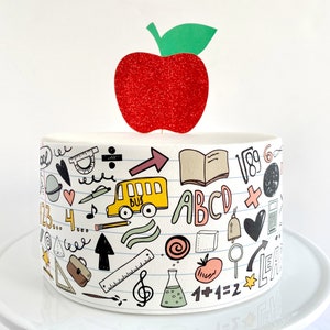 May include: A white cake decorated with a red glitter apple topper and a white and black school-themed design. The design includes a school bus, a book, a pencil, a ruler, a globe, a heart, a music note, a calculator, a bell, a magnifying glass, a test tube, a tomato, and the words "ABC", "1+1=2", and "School".