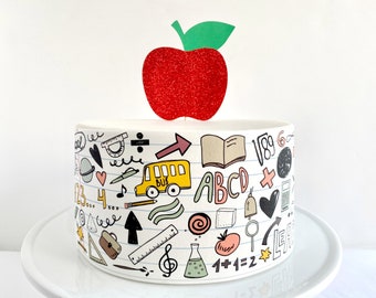 May include: A white cake decorated with a red glitter apple topper and a white and black school-themed design. The design includes a school bus, a book, a pencil, a ruler, a globe, a heart, a music note, a calculator, a bell, a magnifying glass, a test tube, a tomato, and the words "ABC", "1+1=2", and "School".