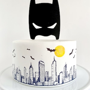 Puede incluir: Un pastel blanco decorado con una silueta negra de Batman y una ciudad con un sol amarillo y murci&eacute;lagos volando en el cielo.