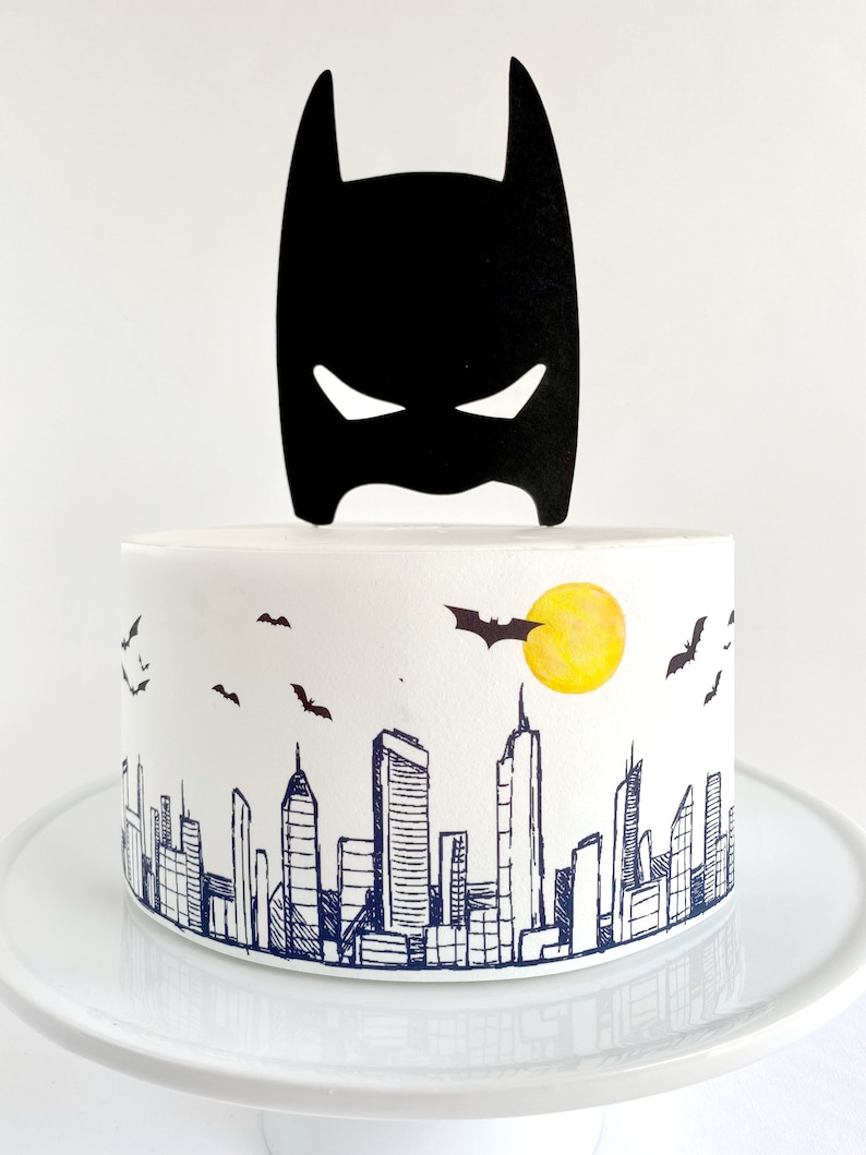 Puede incluir: Un pastel blanco decorado con una silueta negra de Batman y una ciudad con un sol amarillo y murci&eacute;lagos volando en el cielo.