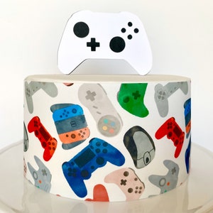 May include: A white cake decorated with a pattern of colorful video game controllers. A white cardboard cutout of a video game controller is on top of the cake.