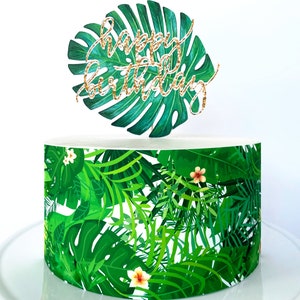 May include: A tropical themed cake decorated with green leaves and flowers. The cake has a topper that says "Happy Birthday" in gold glitter.