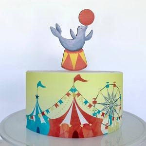 May include: A yellow and blue circus-themed cake with a red and white striped tent, a Ferris wheel, and a gray seal balancing a red ball on its nose.