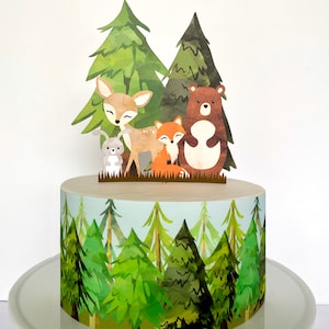 May include: A cake decorated with a woodland theme. The cake topper features a deer, bear, fox, and rabbit. The cake's base is wrapped with a forest scene of green trees against a light blue background.